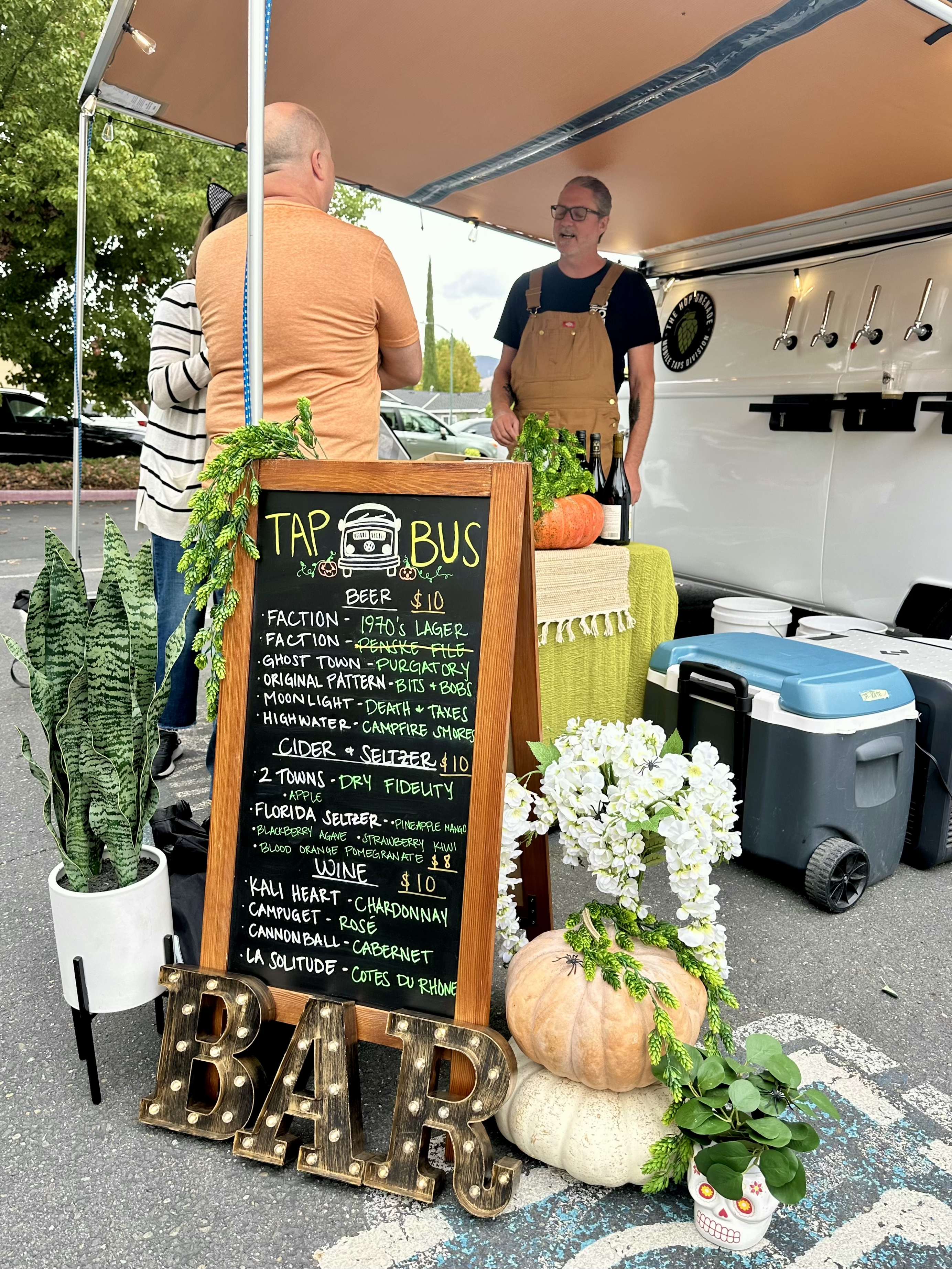 Mobile bar featuring a chalkboard menu, pumpkins, and a bartender serving from a white van.